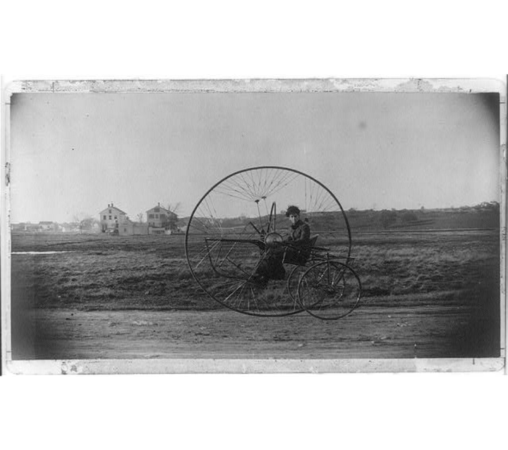 a negative of a very old photograph of a woman in a tricycle with an enormous middle wheel